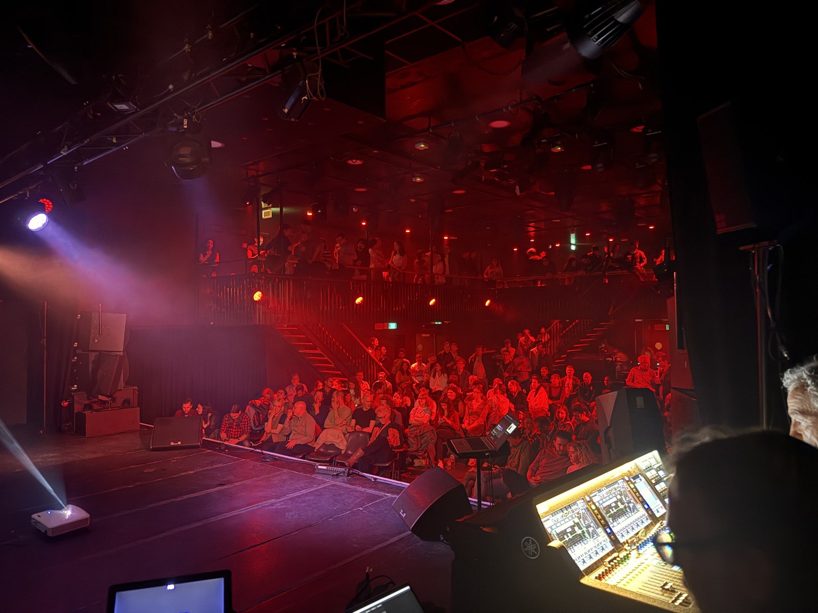 View from behind the soundboard of a full Tolhuistuin auditorium bathed in red light, audience seated on the floor and lining the upper balcony.